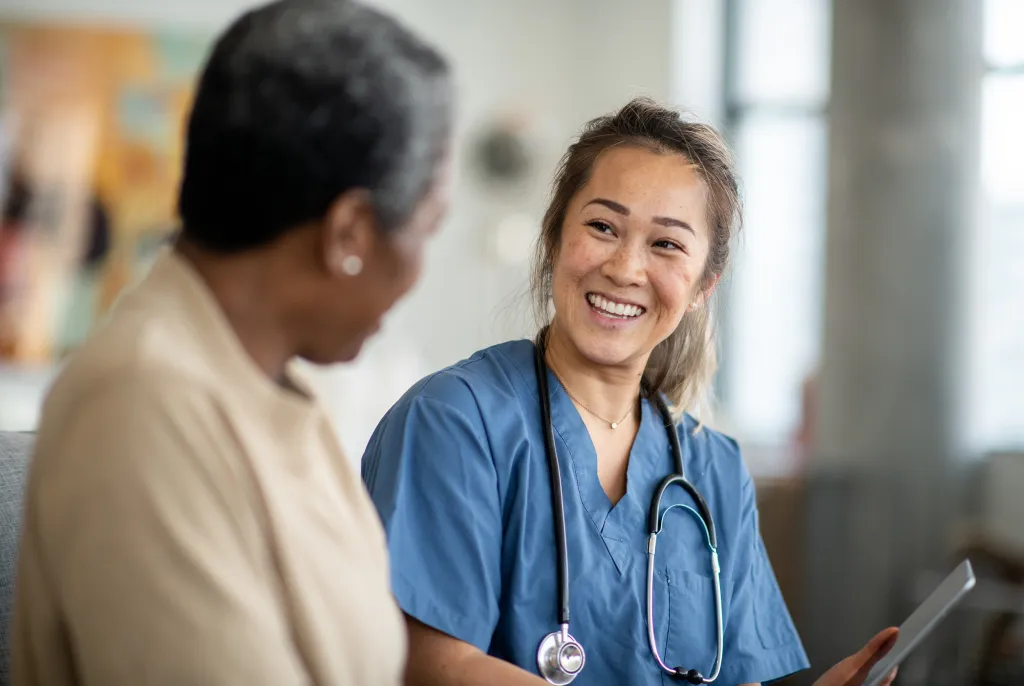 Healthcare professional wearing blue scrubs and a stethoscope sits facing another person in an indoor healthcare setting